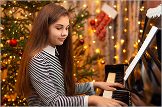 Mädchen mit langen braunen Haaren sitzt am Klavier. Im Hintergrund ein geschmückter Weihnachtsbaum